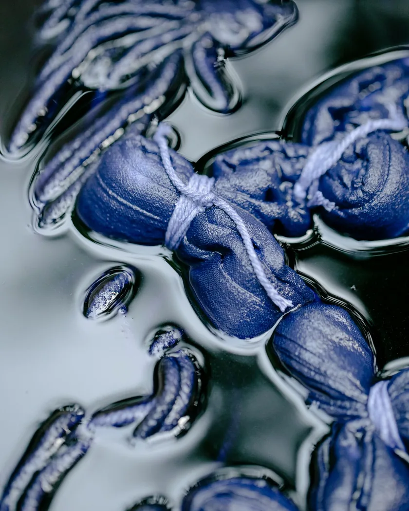 Atados de tela sujetos con hilo y sumergidos en un baño de índigo, con la superficie oscura del líquido cubriendo el tejido azul.